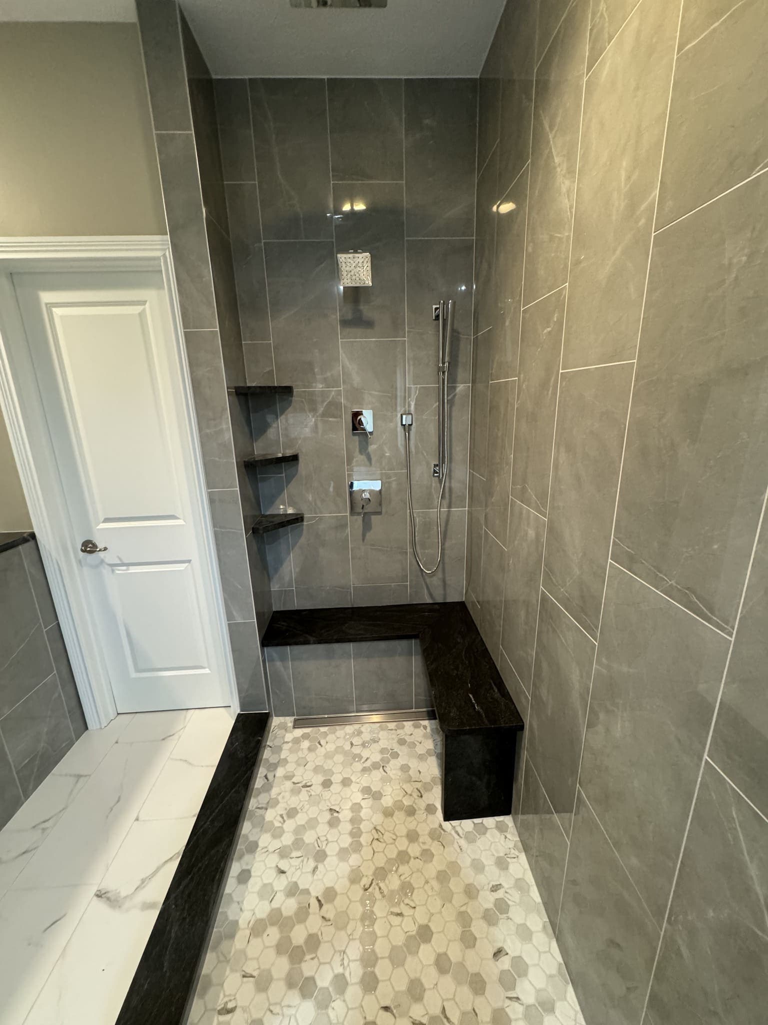 Walk-in shower in large-format grey tile with a dark bench and a hexagon mosaic floor